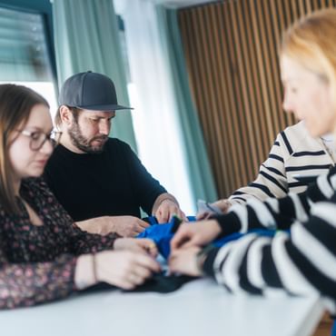 Vier Personen stehen an einem Tisch und halten gemeinsam ein blaues Kleidungsstück in den Händen. Sie schauen konzentriert darauf, während sie es untersuchen oder bearbeiten.