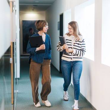 Two women walk side by side through a bright hallway, each holding a laptop or tablet in their arms. They look at each other and smile while talking.