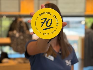 A hand holds a round yellow token displaying “Bründl Sports”, “70” and “Seit 1956” (since 1956). Ski poles and the illuminated interior of a sports shop are visible in the blurred background.