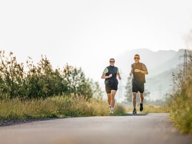 Two people jogging side by side on a tarmac path, surrounded by meadows and trees. In the background, hills and electricity pylons can be seen in the light of the morning sun.