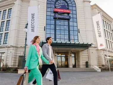 Ein Mann und eine Frau gehen mit Einkaufstaschen an einem großen Gebäude mit dem Schild „McArthurGlen Designer Outlet“ vorbei. Über dem Eingang hängen zwei hohe Banner mit der Aufschrift „Welcome“, und im Hintergrund sind Schaufenster von Geschäften zu sehen.