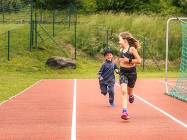 In the picture, two children are running on a track - a girl in front in sportswear, a smaller boy with a cap and tracksuit behind her. Both look at each other with a smile and move towards a football goal at the edge of the track.