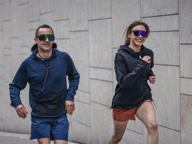 The image shows two people, a man and a woman, jogging together. They are wearing sporty outfits, including hoodies and shorts. Both are wearing striking, mirrored sports sunglasses. The woman is smiling while running. The background features a modern, gray concrete wall with a geometric pattern. The scene conveys a sporty and dynamic atmosphere.