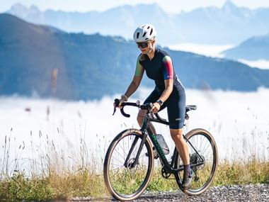 Eine Person mit weißem Helm und dunkler Sportkleidung fährt auf einem schwarzen Fahrrad über einen Schotterweg. Im Hintergrund sind verschwommene Berge und Nebel zu sehen, darüber ein klarer blauer Himmel.