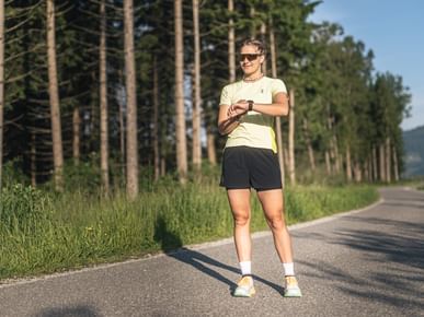 Female runner stands on a paved path at the edge of a forest, looking at her sports watch in sunny conditions.