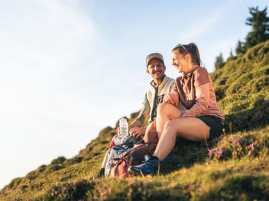 Two people are sitting on a grassy slope, laughing together. Backpacks and a water bottle lie beside them, with hills and trees visible in the warm sunlight in the background.