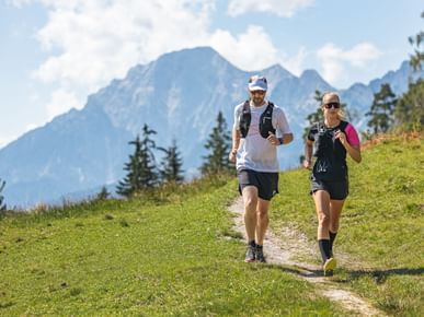 A woman and a man are running side by side on a narrow path through a green meadow. In the background, tall mountains, a few trees, and a sky with white clouds are visible.