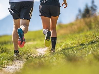Die Beine von zwei laufenden Personen sind von hinten zu sehen, während sie auf einem schmalen Pfad durch grünes Gras laufen. Die Sohlen der Laufschuhe sind sichtbar, und die Umgebung besteht aus einer sonnigen Wiese.