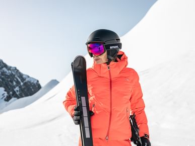 Das Bild zeigt eine Skifahrerin in einer leuchtend orangefarbenen Jacke, die einen Ski hält. Sie trägt einen schwarzen Helm mit einer Skibrille, und im Hintergrund ist eine schneebedeckte Berglandschaft zu sehen.