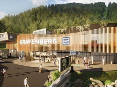 A modern building with a wooden facade stands in front of a forested hillside. The facade shows the large text “GRAFENBERG” and signs reading “Flachau Snow Space Salzburg” and “Bründl Sports,” while several people are walking and standing in front of it.