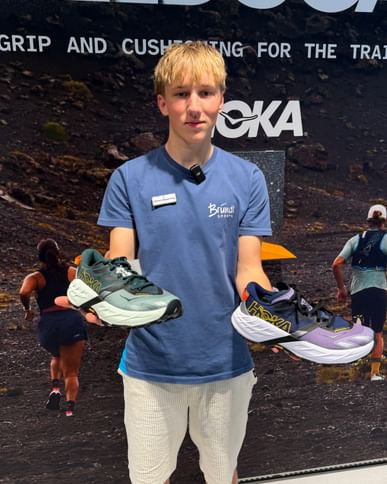 A young man stands in front of a promotional wall, holding a Hoka Speedgoat 7 running shoe in each hand. In the background, there are images of people running and the texts “SPEEDGOAT,” “GRIP AND CUSHIONING FOR THE TRAIL,” and “HOKA” are visible.