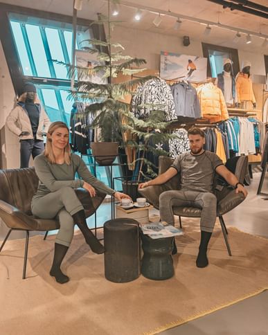 Two adults are sitting in armchairs in a sports shop wearing close-fitting functional underwear. In the background, ski clothing, jackets and mannequins can be seen in a modern salesroom. Between the two of them is a small table with cups.