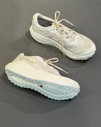 Zwei weiße Sportschuhe liegen auf einem grauen, leicht gesprenkelten Boden. Auf der Seite eines Schuhs ist der Schriftzug „Salomon“ zu lesen, und die Sohlenstruktur ist deutlich sichtbar.