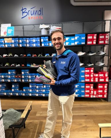 Ein Verkäufer bei Bründl Sports steht in einem Geschäft vor einem Regal mit Sportschuhen und hält zwei Paar Schuhe in den Händen. Er lächelt in die Kamera.