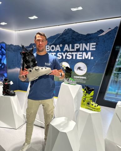 Das Bild zeigt einen Mann in einem Sportgeschäft, der einen Skischuh präsentiert; im Hintergrund sind weitere Skischuhe auf Podesten ausgestellt und eine Wand mit dem Schriftzug "BOA® Alpine Fit System" vor einer Berglandschaft.