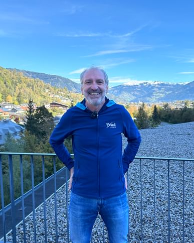 Auf dem Bild steht ein Bründl Sports Mitarbeiter draußen auf einem Balkon oder einer Terrasse, lächelt und trägt eine blaue Bründl Sports-Jacke. Im Hintergrund sind Berge, grüne Hügel und ein blauer Himmel zu sehen.