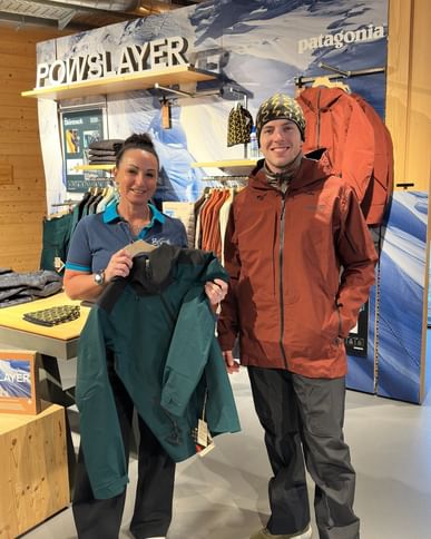 Two people are standing inside a sports retail store in front of a clothing display. The woman is holding a green jacket on a hanger, while the man is wearing a brown outdoor jacket and a beanie. Jackets, shelves, and brand and product signage are visible in the background.