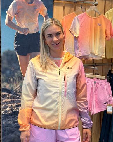 A blonde woman stands in a sportswear store and smiles toward the camera. She is wearing a lightweight jacket in cream, pink, and peach tones with pink shorts. Matching clothing items are displayed on a wooden wall behind her, and a large photo of a runner wearing similar apparel is visible on the left.