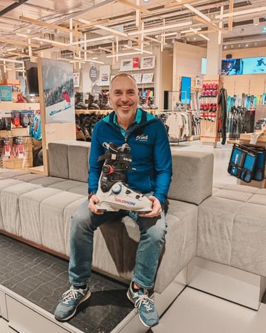 A man is sitting on a bench in a sports shop, holding a pair of Salomon ski boots in his hands. He is wearing casual clothes with a sports jacket and is looking at the camera. In the background, ski boots, skis and sportswear are displayed in a bright, modern salesroom.
