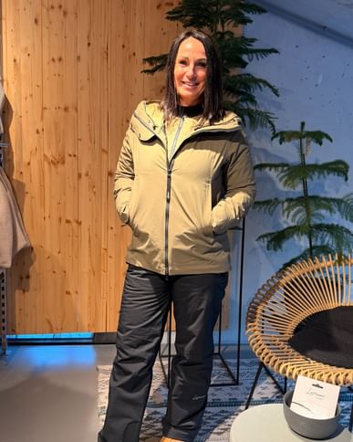 A woman stands in an interior room in front of wood panelling and fir trees, wearing an olive-coloured jacket, black trousers and brown shoes, smiling at the camera.
