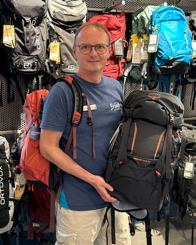 Bründl Sports employee stands with an Osprey Hikelite backpack in front of the backpack wall in the Bründl Sports flagship store