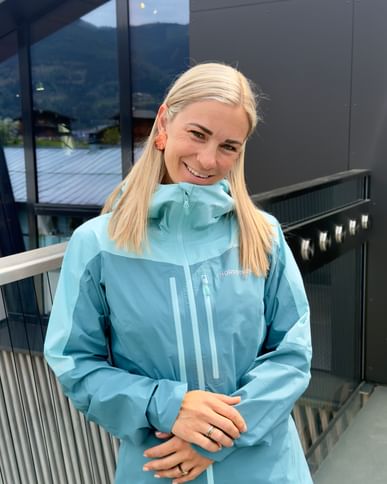 A woman with blonde hair stands on a balcony in front of a modern building with a glass facade, smiling at the camera. She is wearing a light blue Norrona outdoor jacket and pink pants, with mountains and houses visible in the background.
