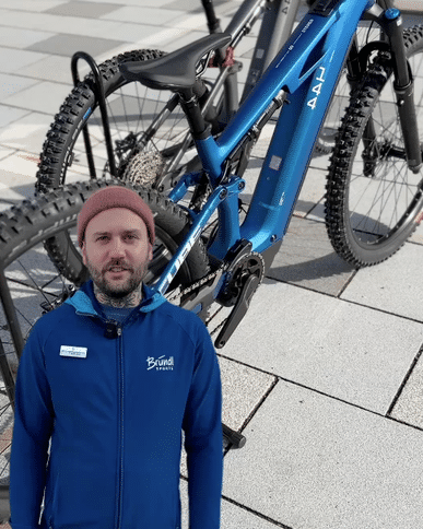 Das Bild zeigt einen Mann in einer blauen Jacke, der vor einem blauen Mountainbike steht