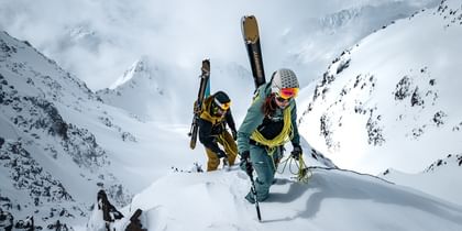 Das Bild zeigt zwei Bergsteiger, die einen schneebedeckten Grat hinaufklettern. Sie tragen Ski auf ihren Rucksäcken und verwenden Kletterausrüstung, um den steilen Anstieg zu bewältigen. Im Hintergrund ist eine beeindruckende, schneebedeckte Berglandschaft zu sehen.