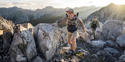 Ein Mann und eine Frau steigen konzentriert durch felsiges Hochgebirgsgelände auf. Beide tragen Dynafit-Bekleidung mit Rucksäcken. Die Sonne bricht durch die Bergkulisse im Hintergrund und verleiht dem Moment eine dynamische Atmosphäre.