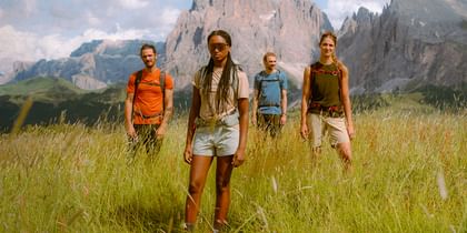 Vier Wanderer stehen selbstbewusst inmitten einer hohen Almwiese mit dramatischem Bergpanorama im Hintergrund. Sie tragen funktionale Outdoorkleidung und schauen entschlossen in Richtung Kamera.
