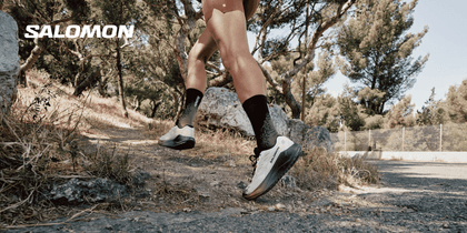 Close-up of a runner’s legs wearing light-coloured Salomon running shoes on a rocky woodland trail. The white “SALOMON” logo appears in the upper left.