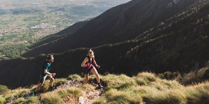 Two trail runners ascend a steep mountain slope with trekking poles, with a breathtaking valley view in the background.