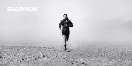 A female trail runner wearing a black jacket, shorts and running vest runs across rocky terrain in the fog. The white “SALOMON” logo appears in the upper left.