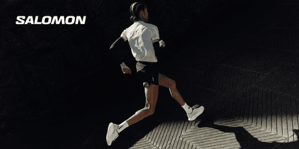 A runner wearing a white shirt, black shorts and light-coloured running shoes runs across dark paving with a geometric pattern. The white “SALOMON” logo appears in the upper left.