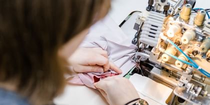 Das Bild zeigt eine Nahaufnahme von jemandem, der an einer Nähmaschine arbeitet und Stoffe zusammennäht. Die Hände der Person führen den Stoff präzise durch die Maschine.
