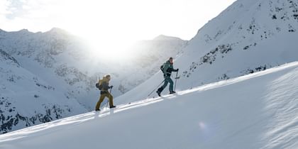 Das Bild zeigt zwei Skifahrer, die mit Tourenski einen verschneiten Berg hinaufsteigen. Die Sonne scheint hinter den Bergen hervor, und die Landschaft ist von schneebedeckten Gipfeln umgeben.