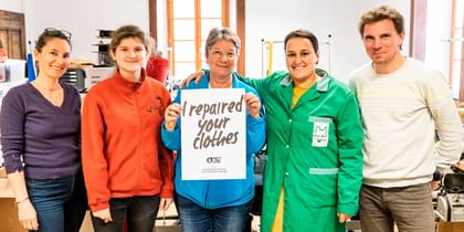Fünf Personen stehen lächelnd in einer Werkstatt, eine Person hält ein Schild mit der Aufschrift „I repaired your clothes“