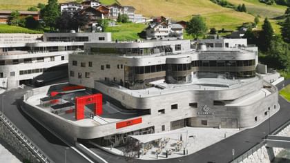 Das Bild zeigt ein modernes, mehrstöckiges Gebäude mit dem Namen "Silvretta Therme Ischgl", umgeben von grünen Hügeln und traditionellen alpenländischen Häusern.