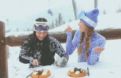 Kari Traa Two women in Kari Traa ski base layers enjoy waffles in the snow at a mountain hut.