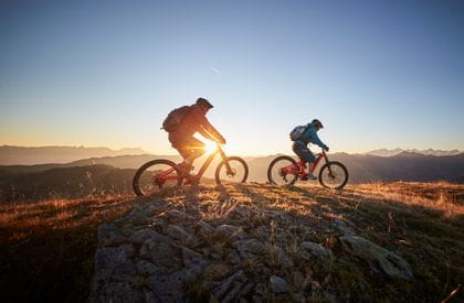 two mountainbikers are riding on a mountain ridge, while the sun sets in the background