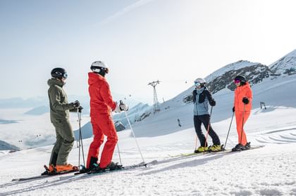 Das Bild zeigt vier Skifahrer, zwei Männer und zwei Frauen, die auf einer Skipiste stehen und sich unterhalten. Sie tragen bunte Skiausrüstungen (grün, rot, orange, blau), umgeben von einer verschneiten Berglandschaft.