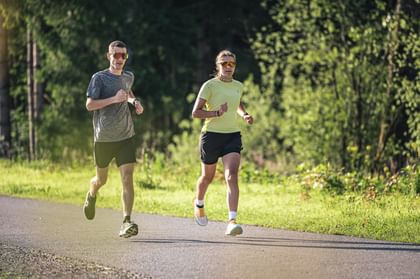 Two runners jog side by side on a paved path through a green forest landscape.