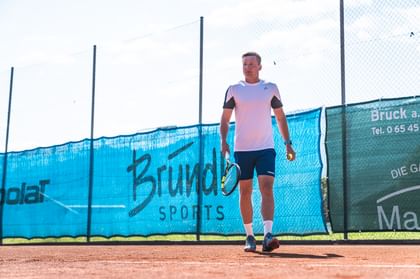Das Bild zeigt einen Tennisspieler auf einem Sandplatz, der mit Schläger und Ball in der Hand bereit für den nächsten Punkt ist. Im Hintergrund ist ein großes Bründl Sports-Banner zu sehen.