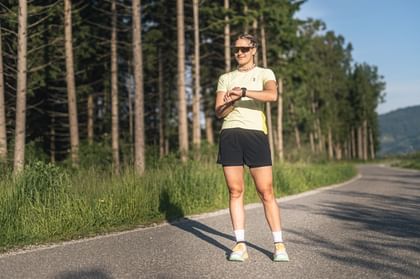 Female runner stands on a paved path at the edge of a forest, looking at her sports watch in sunny conditions.