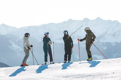 Vier Skifahrer stehen mit Ski und Skistöcken auf einer verschneiten Piste und unterhalten sich. Im Hintergrund sind schneebedeckte Berge zu sehen.
