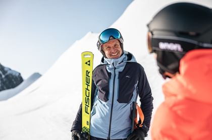 Das Bild zeigt einen Skifahrer in blauer Skijacke, der lächelnd einen gelben Ski trägt. Im Vordergrund sieht man unscharf eine weitere Person in orangefarbener Skibekleidung.