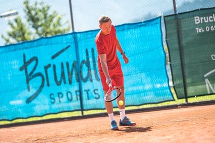 A tennis player in red sportswear plays on a clay court in front of a Bründl Sports advertising banner.