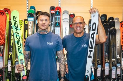 Hans Hofer and Markus Pertl stand in Bründl Sports T-shirts in front of a wall of skis, each holding a pair of Fischer and Head skis in their hands.