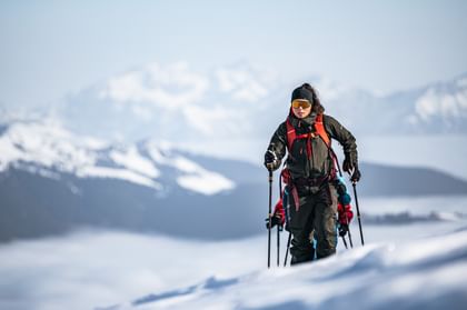 Eine Person in Skitouren-Ausrüstung steigt einen schneebedeckten Hang hinauf, während im Hintergrund eine beeindruckende Berglandschaft mit wolkenverhangenen Tälern zu sehen ist.