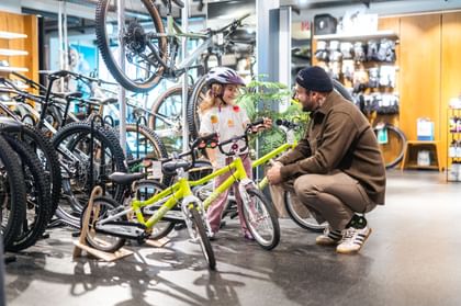Das Bild zeigt ein kleines Mädchen, das in der Bründl Sports Bikeworld mit einem Erwachsenen ein Kinderfahrrad betrachtet, während sie fröhlich miteinander sprechen.